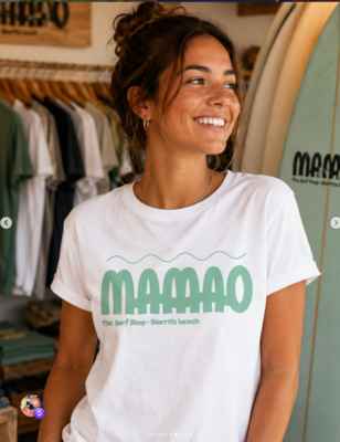 Jeune femme souriante portant un t-shirt blanc floqué du logo vert Mamao The Surf Shop dans une boutique de surf